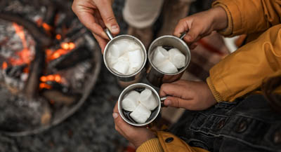 People holding mugs of cocoa with marshmallows around campfire.